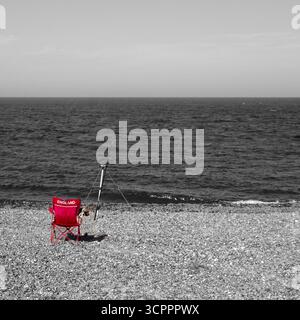 Schwarz-weiß-Aufnahme eines Mannes beim Angeln am Strand mit einem Stuhl in Rot Stockfoto