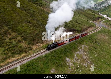 Aus der Vogelperspektive einer alten Dampfeisenbahn, die sich durch die grünen, steilen Hänge schlängelt, mit weißen Rauchschwaden im Kontrast zur grünen Landschaft, Furka Pass, Schweiz. Stockfoto