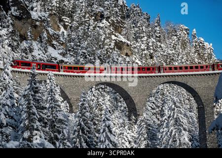 Aus der Vogelperspektive überquert ein pulsierender roter Zug eine schneebedeckte Steinbogenbrücke inmitten der mattierten immergrünen und felsigen Klippen in Filisur, Schweiz. Stockfoto