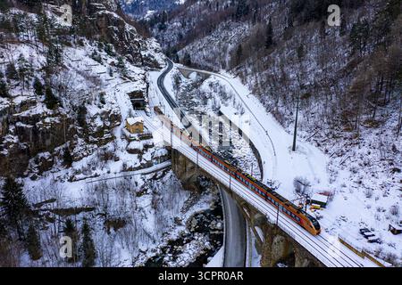 Aus der Vogelperspektive eines pulsierenden roten Zuges, der eine Steinbrücke über einen Gletscherfluss durchquert, inmitten von schneebedeckten Wäldern und Bergen, Filisur, Schweiz. Stockfoto