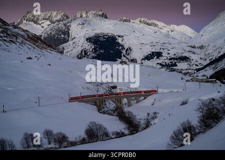 Aus der Vogelperspektive eines pulsierenden roten Zuges, der eine Steinbrücke in einem schneebedeckten Tal durchquert, eingerahmt von majestätischen, schneebedeckten Bergen, Filisur, Schweiz Stockfoto