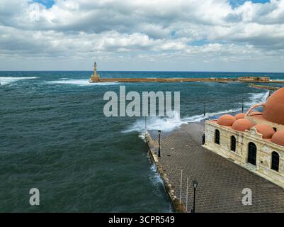 Aus der Vogelperspektive der Moschee von Kioutsouk Hassan Kuppeln im Kontrast zum turbulenten Meer und dem fernen Leuchtturm Chania, Chania, Griechenland. Stockfoto