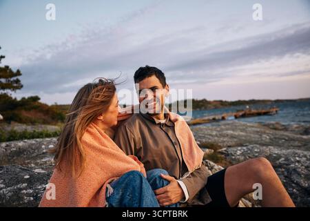 Lächelndes Paar, das in der Abenddämmerung auf einem Felsen sitzt, in einer Decke in der Nähe des Meeres Stockfoto