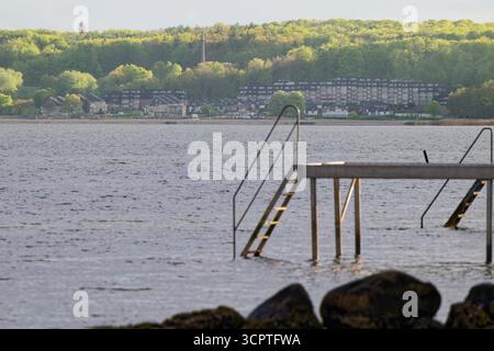 Ein stabiler Metallpfeiler erstreckt sich über das Wasser, wo zwei Treppen ins Meer führen, während hinter den Geländern ein Strand und mehrere Häuser eingebettet sind Stockfoto