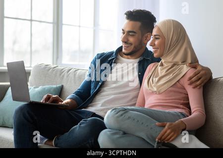 Entspanntes Paar aus dem Nahen Osten sitzt auf der Couch und benutzt einen Laptop Stockfoto