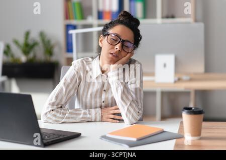 Müde Geschäftsfrau schläft am Arbeitsplatz, während sie an einem Laptop-Computer in einem modernen Bürogebäude arbeitet Stockfoto