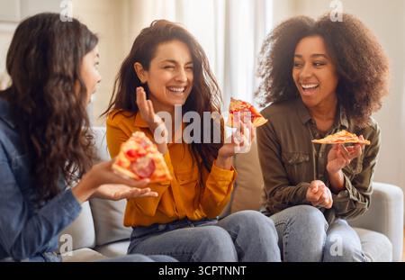 Drei verschiedene Freundinnen genießen Pizza und amüsieren sich drinnen Stockfoto