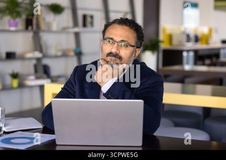 Nachdenklicher indischer Geschäftsmann in Anzug sitzt am Schreibtisch mit Laptop Stockfoto