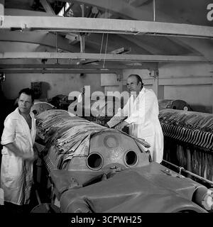 Arbeiter in weißen Mänteln betreiben in den 1970er Jahren große industrielle Pressen in einer Werkstatt des Slawjansk Fittings and Isolator Plant (AIZ) in der ukrainischen SSR Dieses einfarbige Archivfoto erfasst einen Herstellungsprozess in der sowjetischen Fabrik, der möglicherweise das Pressen von Verbundwerkstoffen oder Gewebematerialien beinhaltet. Die Szene zeigt die Handarbeit und die schweren Maschinen, die bei der Herstellung spezieller Komponenten in der industriellen Donbass-Region beteiligt sind Stockfoto