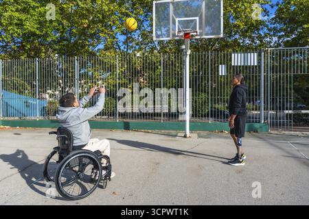 Zwei behinderte Freunde spielen im Freien Basketball, einer schießt aus einem Rollstuhl, der andere mit einer Beinprothese verteidigt sich und teilt adaptive s Stockfoto