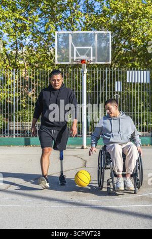 Zwei Freunde spielen Basketball auf einem Platz im Freien, ein Mann mit einer Beinprothese, der andere im Rollstuhl dribbelt einen gelben Ball und feiert Incluusi Stockfoto