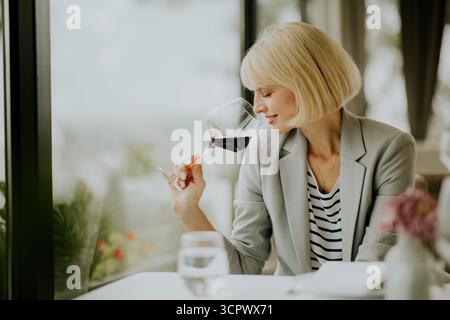Eine junge Frau in einem grauen Blazer sitzt an einem Café-Tisch und isst Rotwein. Sie blickt aus dem Fenster, in Gedanken verloren, während Blumen den Tisch schmücken. Das A Stockfoto