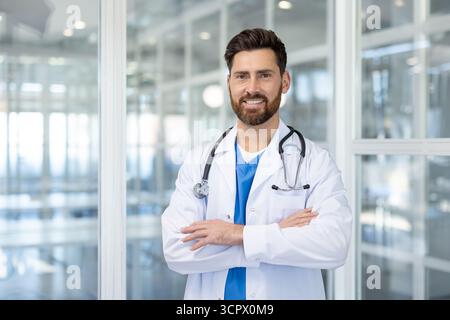 Professioneller männlicher Arzt mit Bart in weißem Labormantel und Stethoskop, selbstbewusst mit gekreuzten Armen in einem hell beleuchteten modernen Krankenhauskorridor stehend Stockfoto