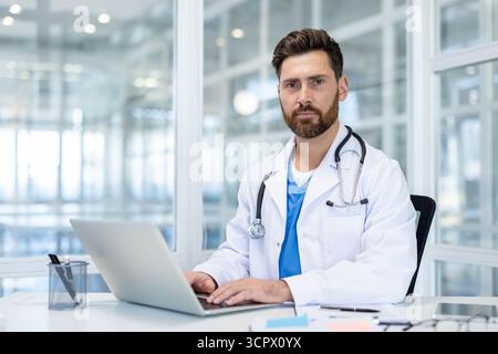 Selbstbewusster männlicher Arzt, der an seinem Schreibtisch an einem Laptop arbeitet, einen weißen Labormantel und ein Stethoskop trägt und für Gesundheit, Telemedizin und medizinische Professionalität steht Stockfoto