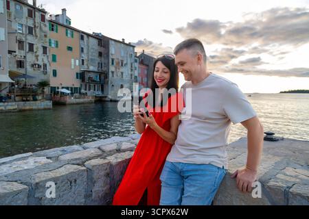 Glückliches Paar mit Smartphone am Ufer von Rovinj. Ein lächelndes Paar lehnt sich auf eine Steinmauer am Meer in Rovinj, Kroatien, und schaut auf ein Smartphone Stockfoto