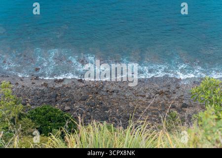 Hochwinkelblick von einer steilen Klippe mit einem dunklen felsigen Strand und schaumigen weißen Wellen des tiefblauen tropischen Ozeans. Stockfoto