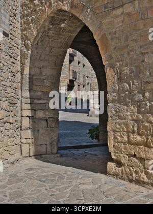 Mittelalterlicher Steinbogen in der Village Street - Eine sonnendurchflutete, bogenförmige Steinpassage, die in einen historischen, ruhigen Stadtplatz mit traditionellen Steinhäusern führt Stockfoto