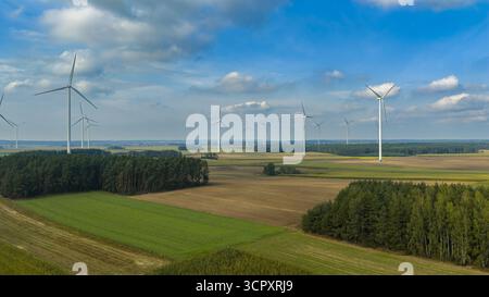 Panoramablick auf Windturbinen in offenen Feldern, umgeben von grünen und braunen landwirtschaftlichen Flächen, dichten Baumgruppen und einem teilweise bewölkten Himmel. Stockfoto