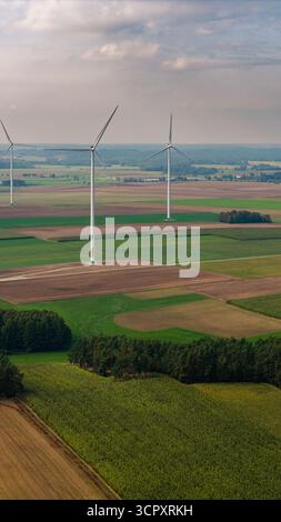 Panoramablick auf Windturbinen in einer ländlichen Landschaft mit grünen und braunen Feldern, Bäumen und einem teilweise bewölkten Himmel am Horizont. Stockfoto