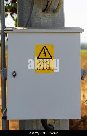Ein grauer Schaltkasten, der auf einem Metallmast montiert ist, mit einem gelben Warnschild mit einem Blitzsymbol und einem Text in türkischer Sprache mit der Aufschrift „Gefahr von“ Stockfoto
