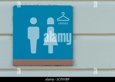 Das Bild zeigt ein blaues quadratisches Schild mit weißen Unisex-Figuren und einem Kleiderbügelsymbol, das an einer weißen Holzwand befestigt ist. Stockfoto