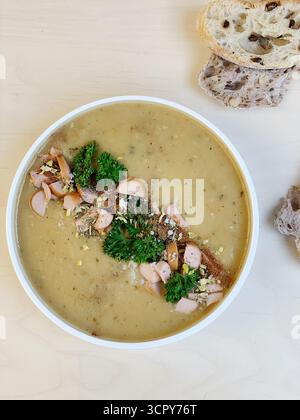 Ein Schuss über der Decke einer herzhaften Schüssel mit traditioneller Linsensuppe in einer weißen Schüssel. Die dicke, cremige, blassgrüne Suppe wird mit Würstchen und Kräutern garniert Stockfoto