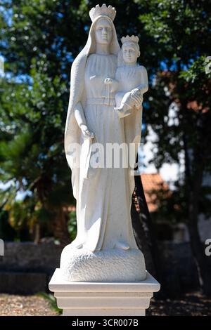 Eine heitere Statue aus weißem Stein der Jungfrau Maria, die das Kind in der Hand hält, gekrönt auf einem Podest in einem sonnendurchfluteten Garten steht. Perfekt für religiöse, Gedenkstätten oder Stockfoto