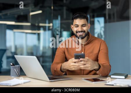 Ein junger indischer Geschäftsmann sitzt an seinem Schreibtisch in einem modernen Büro und navigiert gerne auf seinem Smartphone und repräsentiert Konzepte wie mobile Kommunikation, Unternehmenstechnologie und Konnektivität Stockfoto