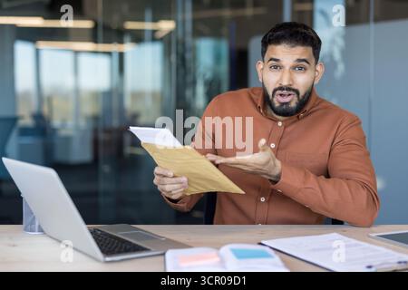 Junger Geschäftsmann, der am Schreibtisch im Büro sitzt und überraschende Neuigkeiten erhält, während er ein wichtiges Dokument aus einem braunen Umschlag öffnet und mit einem schockierten Ausdruck reagiert Stockfoto