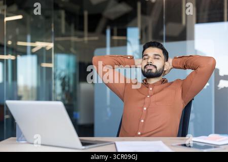 Junger bärtiger Mann in einem braunen Hemd, der sich in einem Bürostuhl mit Händen hinter dem Kopf und geschlossenen Augen entspannt und mit einem Laptop an seinem Schreibtisch eine Pause von der Arbeit macht Stockfoto
