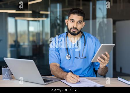 Männlicher Arzt in blauen Peelings und Stethoskop sitzt am Schreibtisch mit Tablet und Laptop und schreibt Notizen-fokussiertes Porträt, das moderne Telemedizin, digitale Dokumentation und klinische Versorgung vermittelt Stockfoto