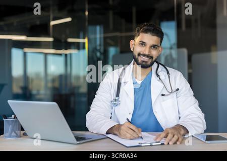 Junge indische Ärztin im weißen Mantel mit Stethoskop lächelt an den Schreibtisch, schreibt auf einem Klemmbrett neben Laptop und Tablet, vermittelt moderne Telemedizin und professionelle Pflege Stockfoto