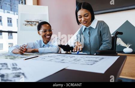 Zwei junge Frauen, eine davon im Rollstuhl, arbeiten an architektonischen Entwürfen und Entwürfen in einem Büro zusammen, das Kreativität und Vielfalt zum Ausdruck bringt. Stockfoto