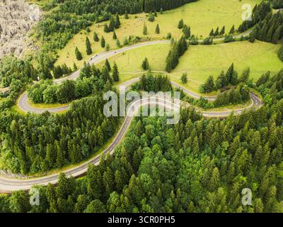 Dieser Blick aus der Vogelperspektive fängt die Serpentinenstraßen der Alpen ein, umgeben von dichten Wäldern. Die Kurven sind ein einladender Weg für Reisende, die die landschaftliche Schönheit auf einem Roadtrip erkunden. Stockfoto