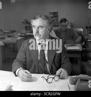 Ein nachdenklicher Manager mittleren Alters oder leitender Ingenieur mit Schnurrbart hält um 1985 an seinem überdachten Schreibtisch in einem geschäftigen Büro der Slawjansk Armature and Insulator Plant (AIZ), ukrainische SSR, inne. Dieses authentische Schwarzweiß-Vintage-Foto fängt die tägliche Atmosphäre eines sowjetischen Arbeitsplatzes ein. Es zeigt einen Verwaltungsexperten mit Papierkram und einem Drehtelefon und symbolisiert die intellektuelle Entscheidungsarbeit, die die Industrie der friedlichen Donbass-Region geleitet hat Stockfoto
