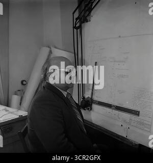 Ein leitender Ingenieur oder Architekt sitzt nachdenklich an seinem professionellen Zeichentisch und untersucht einen technischen Entwurf im Konstruktionsbüro der Slavjansk Armature and Insulator Plant (AIZ), ukrainische SSR, um 1985. Dieses klassische Schwarz-weiß-Porträt fängt das Wesen der intellektuellen Arbeit in der Zeit vor dem Computer der sowjetischen Industrie ein und symbolisiert das akribische Planungs- und Ingenieurwissen, das die Grundlage für die gesamte Produktion in der friedlichen Donbass-Region bildete. Stockfoto