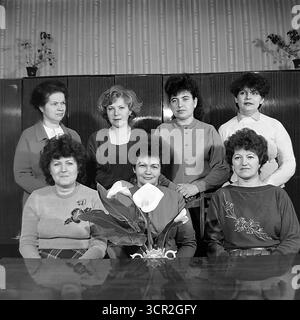 Ein formelles Gruppenporträt eines weiblichen Arbeitskollektivs, wahrscheinlich einer Verwaltungs- oder Ingenieurabteilung, das sich um 1985 in einem Büro im Slawjansk Armature and Insulator Plant (AIZ), ukrainische SSR, posierte. Dieses klassische Schwarz-weiß-Vintage-Foto mit seinen charakteristischen Frisuren und Mode aus den 1980er Jahren fängt die Kameradschaft und den Stolz sowjetischer Frauen am Arbeitsplatz ein und symbolisiert die starken sozialen Bindungen und das friedliche Alltagsleben im Donbass. Stockfoto