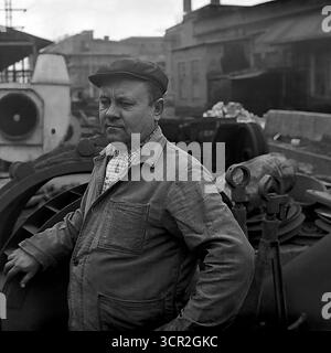 Ein selbstbewusstes Außenporträt eines erfahrenen Fabrikarbeiters mittleren Alters in Kappe und Arbeitsjacke, der um 1985 auf dem Industriehof eines Werks in Sloviansk, ukrainische SSR, posierte. Dieses authentische Schwarz-weiß-Foto fängt den Charakter eines erfahrenen Profis ein, eines Meisters seines Handwerks, dessen beständiger Blick den kompetenten und zuverlässigen Kern der sowjetischen Industriearbeit verkörpert und den produktiven Geist des friedlichen Donbas symbolisiert. Stockfoto