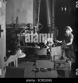 Eine junge Arbeiterin, die vor Stolz und Konzentration lächelt, betreibt eine halbautomatische Produktionslinie für Keramikisolatoren in einer dramatisch beleuchteten Werkstatt in der Slavjansk Armature and Insulator Plant (AIZ), etwa 1985. Dieses stimmungsvolle Schwarzweiß-Foto aus Sloviansk, der ukrainischen SSR, fängt die Synergie zwischen Mensch und Maschine in der sowjetischen Industrie ein, hebt die wesentliche Rolle der qualifizierten weiblichen Arbeit hervor und symbolisiert den zielgerichteten, kreativen Geist des friedlichen Donbass. Stockfoto