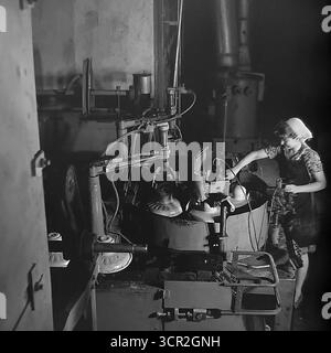 Eine junge Arbeiterin, die vor Stolz und Konzentration lächelt, betreibt eine halbautomatische Produktionslinie für Keramikisolatoren in einer dramatisch beleuchteten Werkstatt in der Slavjansk Armature and Insulator Plant (AIZ), etwa 1985. Dieses stimmungsvolle Schwarzweiß-Foto aus Sloviansk, der ukrainischen SSR, fängt die Synergie zwischen Mensch und Maschine in der sowjetischen Industrie ein, hebt die wesentliche Rolle der qualifizierten weiblichen Arbeit hervor und symbolisiert den zielgerichteten, kreativen Geist des friedlichen Donbass. Stockfoto