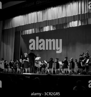 Ein Sinfonieorchester unter der Leitung eines Dirigenten und mit einer Solistin spielt um 1985 ein klassisches Musikkonzert auf der Bühne eines Kulturpalastes in Sloviansk, Ukrainische SSR. Dieses stimmungsvolle Schwarzweiß-Foto aus der Perspektive des Publikums zeigt ein bedeutendes kulturelles Ereignis in einer sowjetischen Industriestadt mit einem patriotischen Slogan, der über der Bühne sichtbar ist und die Förderung der hohen Kunst und das reiche kulturelle Leben im friedlichen Donbass symbolisiert Stockfoto