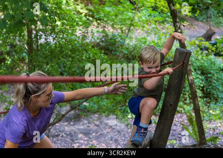 Kinder spielen auf Rutsche und Kletterseilen auf einem von Bäumen umgebenen Naturspielplatz. Stockfoto