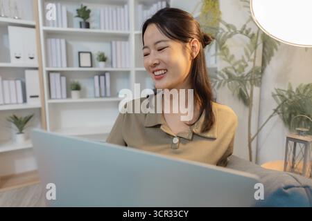 Eine junge Frau sitzt auf einem Sofa mit einem Laptop und lächelt, während sie auf den Bildschirm blickt. Sie wirkt entspannt und engagiert, genießt Online-Kommunikation, fernab Stockfoto