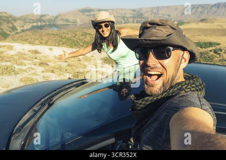 Glückliches junges weißes Paar, das Selfie macht, während es in einem schwarzen Mietwagen sitzt und eine Roadtrip durch Europa macht. Outdoor-Lifestyle-Aktivität auf Roadtrip Stockfoto