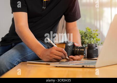Zugeschnittene Aufnahme eines jungen, fröhlichen Fotografen Mann, der Fotokamera benutzt, während er in seinem Büro arbeitet. Stockfoto