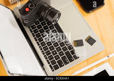 Zugeschnittenes Bild eines Mobiltelefons, eines Laptops, eines Z-Stifts und eines Notizblocks mit Flash-Laufwerken und einer Kamera auf einem Holztisch. Gegenstände des Fotografen im Arbeitsauftrag Stockfoto