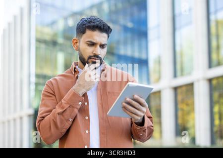 Junger Geschäftsmann, der draußen in einer Stadt mit einem digitalen Tablet steht, fokussiert und nachdenklich ist, während er unterwegs Daten analysiert, Strategien plant und Lösungen erforscht Stockfoto