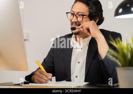 Seitenansicht des Callcenters für asiatische Männer, Kundenservice, Telesales in formellem Anzug, das Headset oder Kopfhörer trägt und mit dem Kunden in einem modernen Büro spricht. Stockfoto