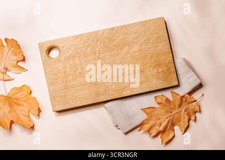 Leeres Schneidebrett auf beigefarbenem Hintergrund mit herbstgelben Blättern, saisonales Essensmockup. Flache Lagen Stockfoto