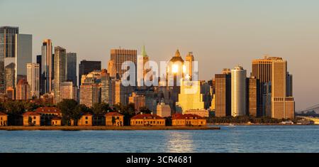 Blick auf die Skyline von Lower Manhattan bei Sonnenuntergang mit den Wolkenkratzern des Finanzviertels. Ellis Island historische Gebäude im Vordergrund Stockfoto
